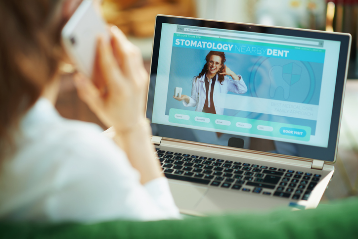 Seen from behind woman at home in sunny day using a mobile phone and looking at digital dental site on a laptop while sitting on a couch.